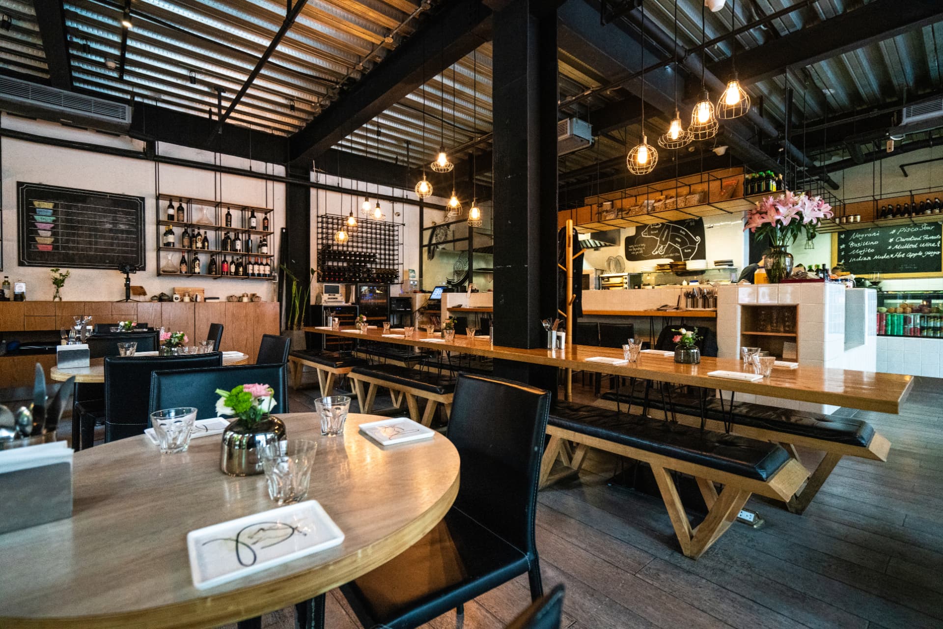 Warm Rivulet restaurant interior with communal timber tables, black steel columns, pendant lighting, and an open kitchen counter